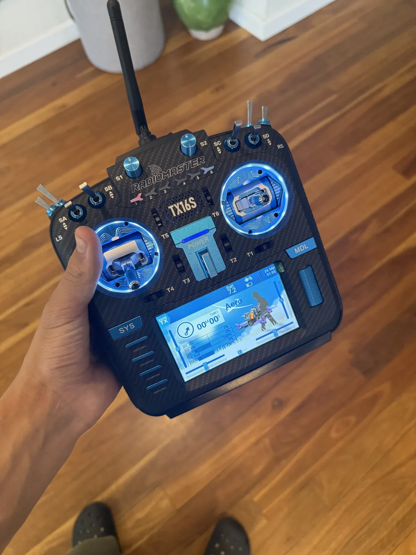 A RadioMaster TX16S radio controller held in one hand, with blue gimbals and its screen lit up.