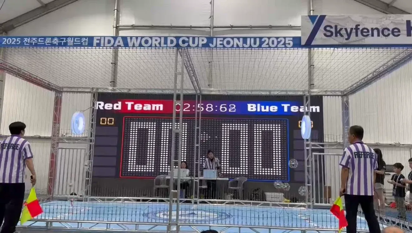 The drone soccer arena at the FIDA World Cup Jeonju 2025, a netted cage with a red team versus blue team scoreboard and two referees.