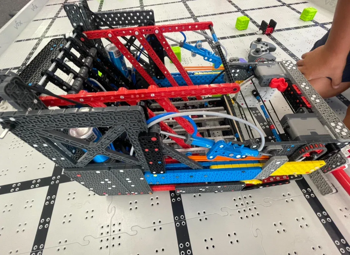 A VEX IQ robot with red and black beams on a Full Volume field, with green blocks behind it.