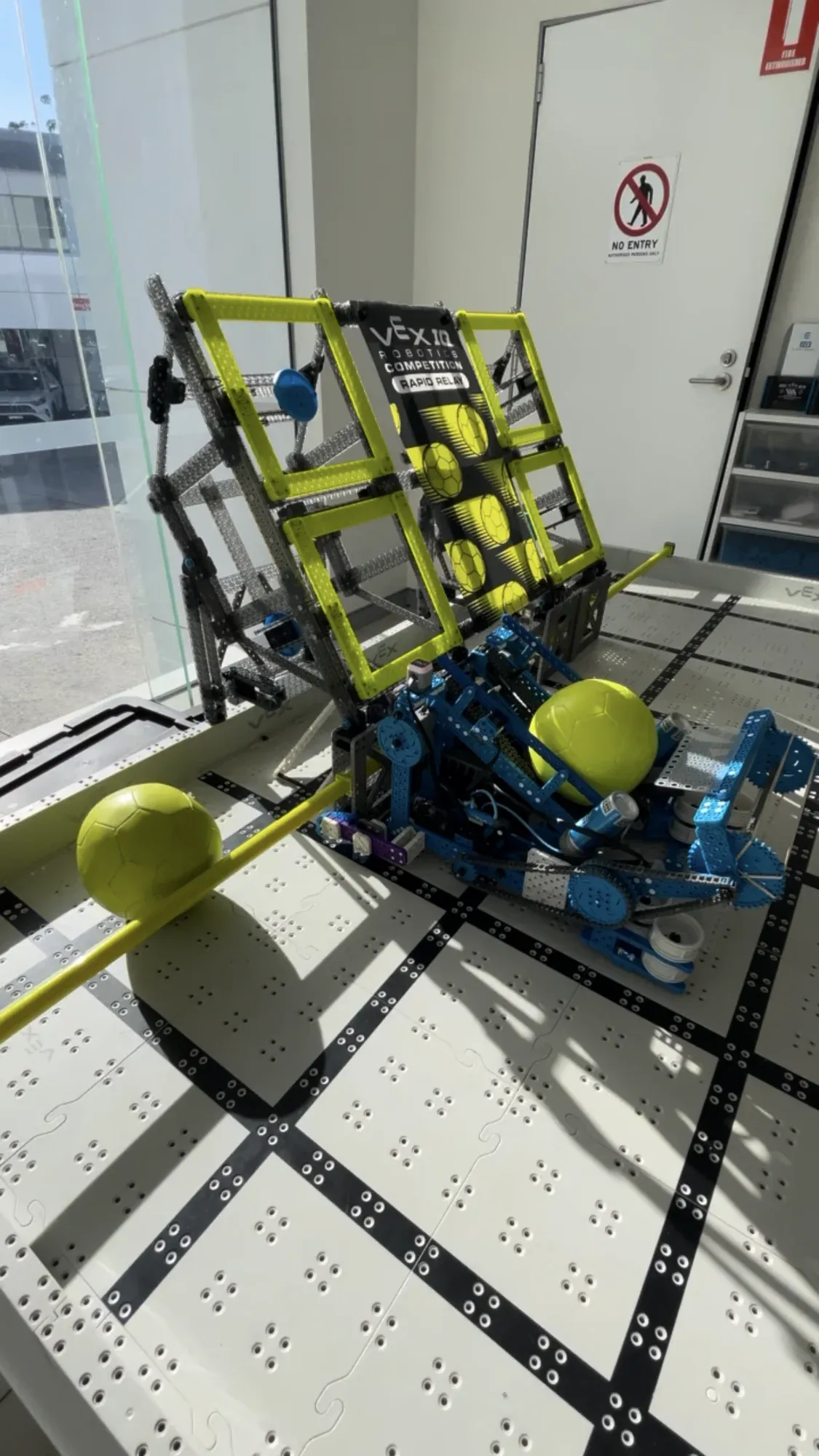 A blue VEX IQ robot on the Rapid Relay field beside the yellow goal, with yellow balls around it.