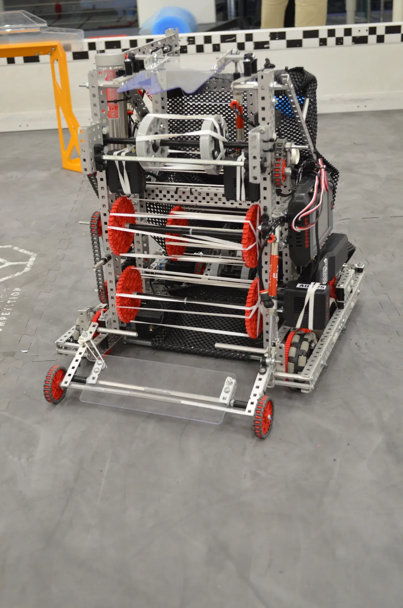 A VEX V5 robot with red wheels and a white intake, sitting on a grey floor.