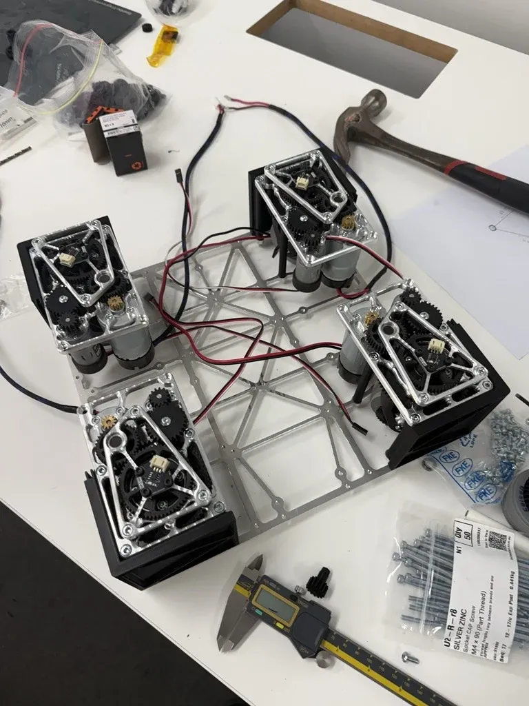 Four swerve modules bolted to a small aluminium plate on a workbench, with wiring between them.